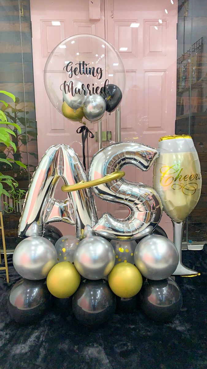Decorative balloon arrangement with 'Getting Married' and 'AS' balloons, a glass of champagne, and a pink wall in the background.
