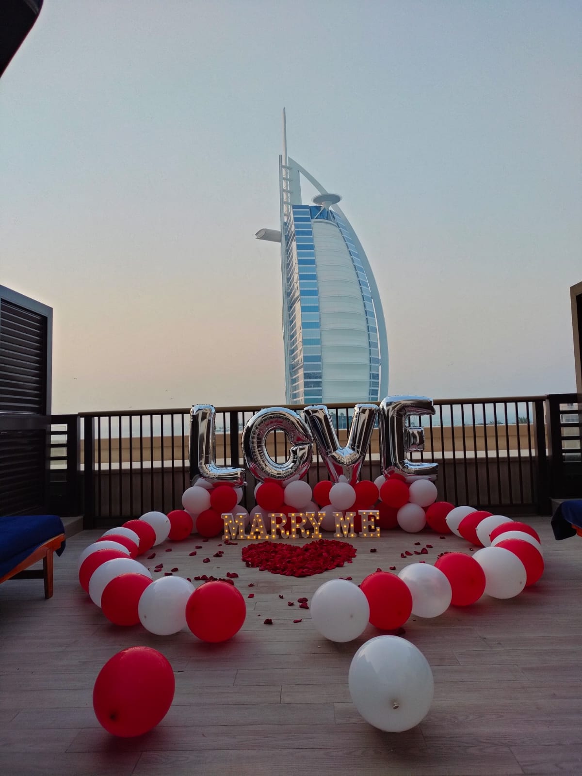 Decorative setup with balloons and 'LOVE ME' sign on a rooftop with a cityscape background