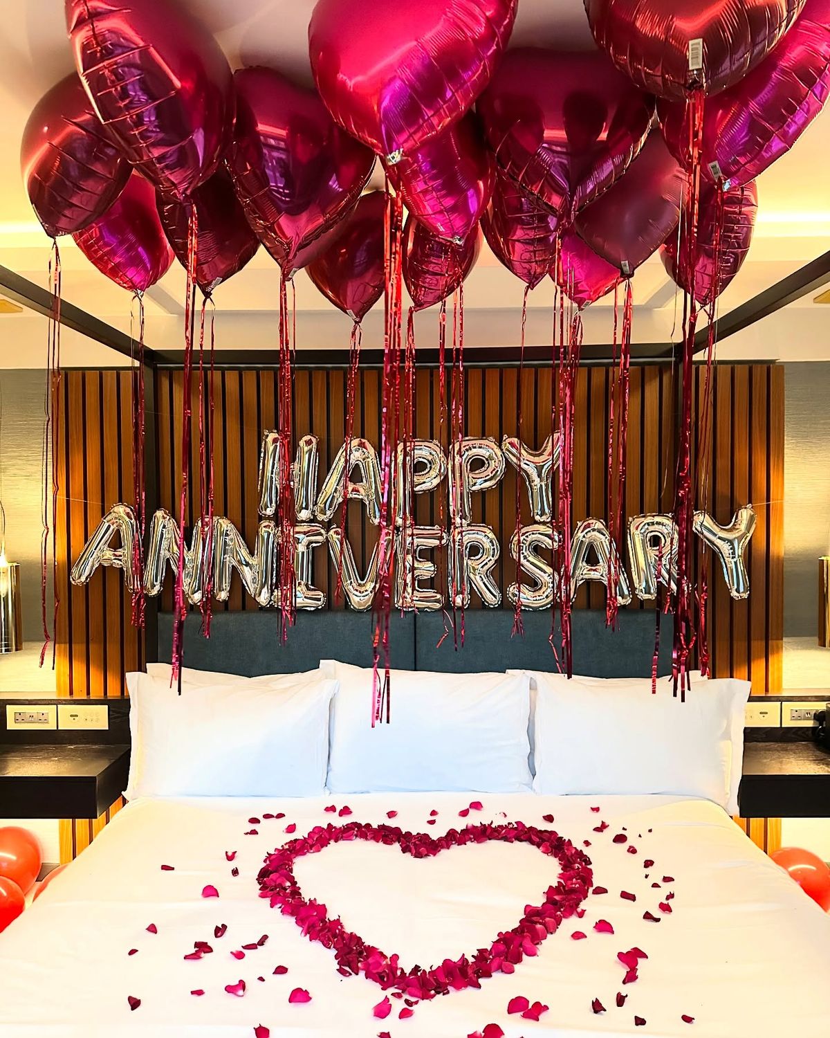 Decorated bedroom with 'Happy Anniversary' balloons and heart-shaped rose petals on a bed.