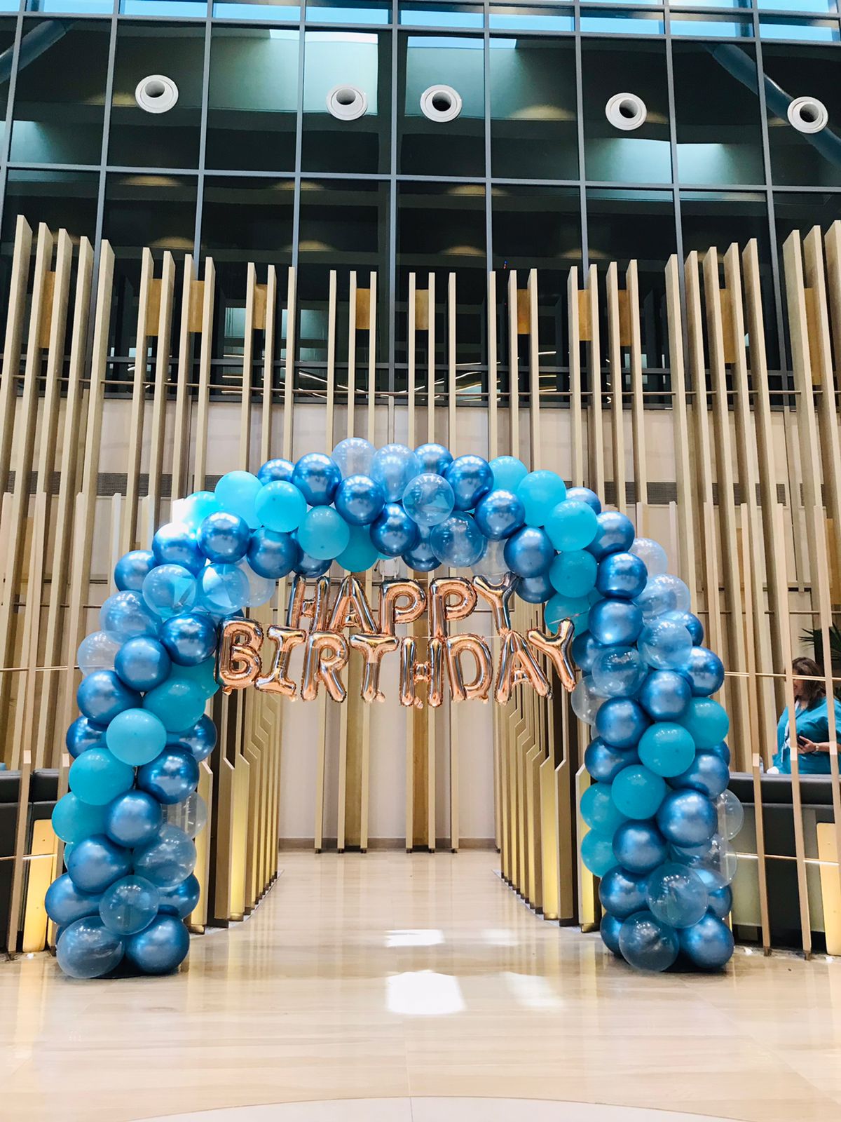 Blue balloon arch with 'Happy Birthday' sign in front of a modern building entrance.