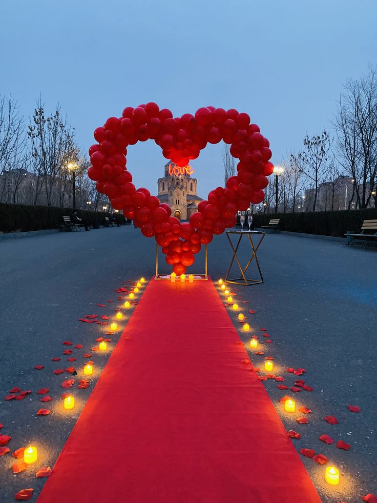 Heart-shaped arrangement of red balloons on a red carpet with candles, set against a cityscape.