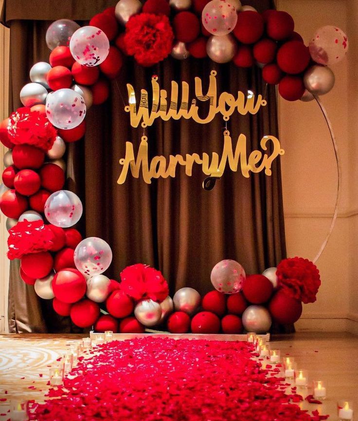 Decorative setup with balloons, flowers, and 'Will You Marry Me?' sign on a brown curtain background.